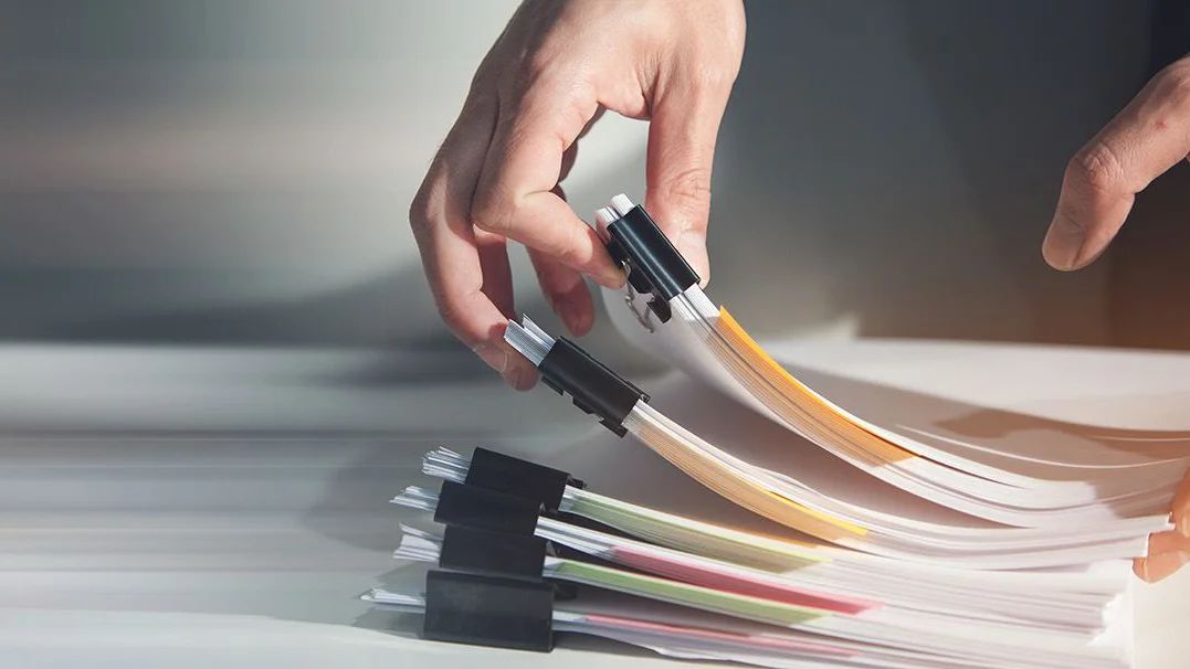 A hand flipping through stacks of paper, each stack held together by binder clips.