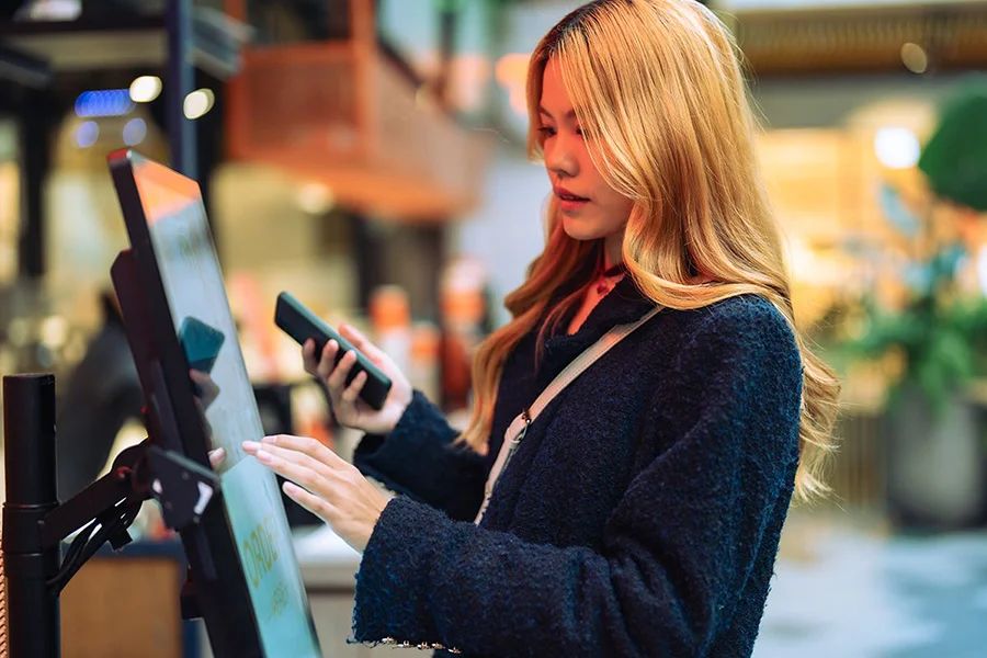 A young woman holding her phone while using a self-service kiosk to process a return at a retail store.